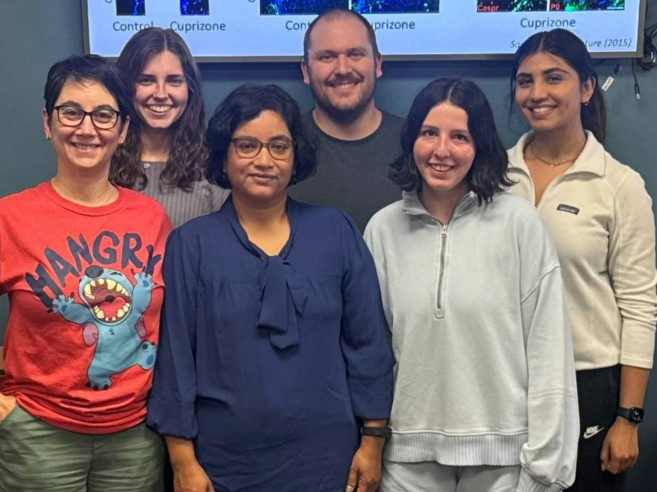 A group of six individuals stands together in a casual setting, displaying a mix of attire and smiling in front of a presentation screen.